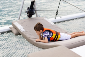 Child wearing a life jacket relaxing on the front deck netting of a family yacht charter